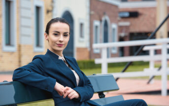 A confident person wearing a navy blue suit is sitting on a bench outdoors. They have short dark hair tied back and are smiling slightly. The background features buildings and a grassy area, suggesting an urban or suburban setting.