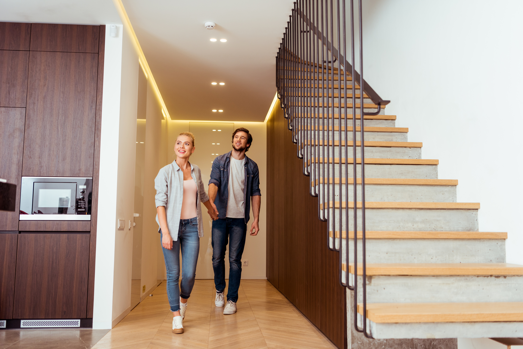 A smiling couple walks hand-in-hand through a hallway of a modern home. Wooden cabinets and an integrated microwave are on the left, while a staircase with metal railings ascends to the right. The hallway is well-lit with recessed lighting.