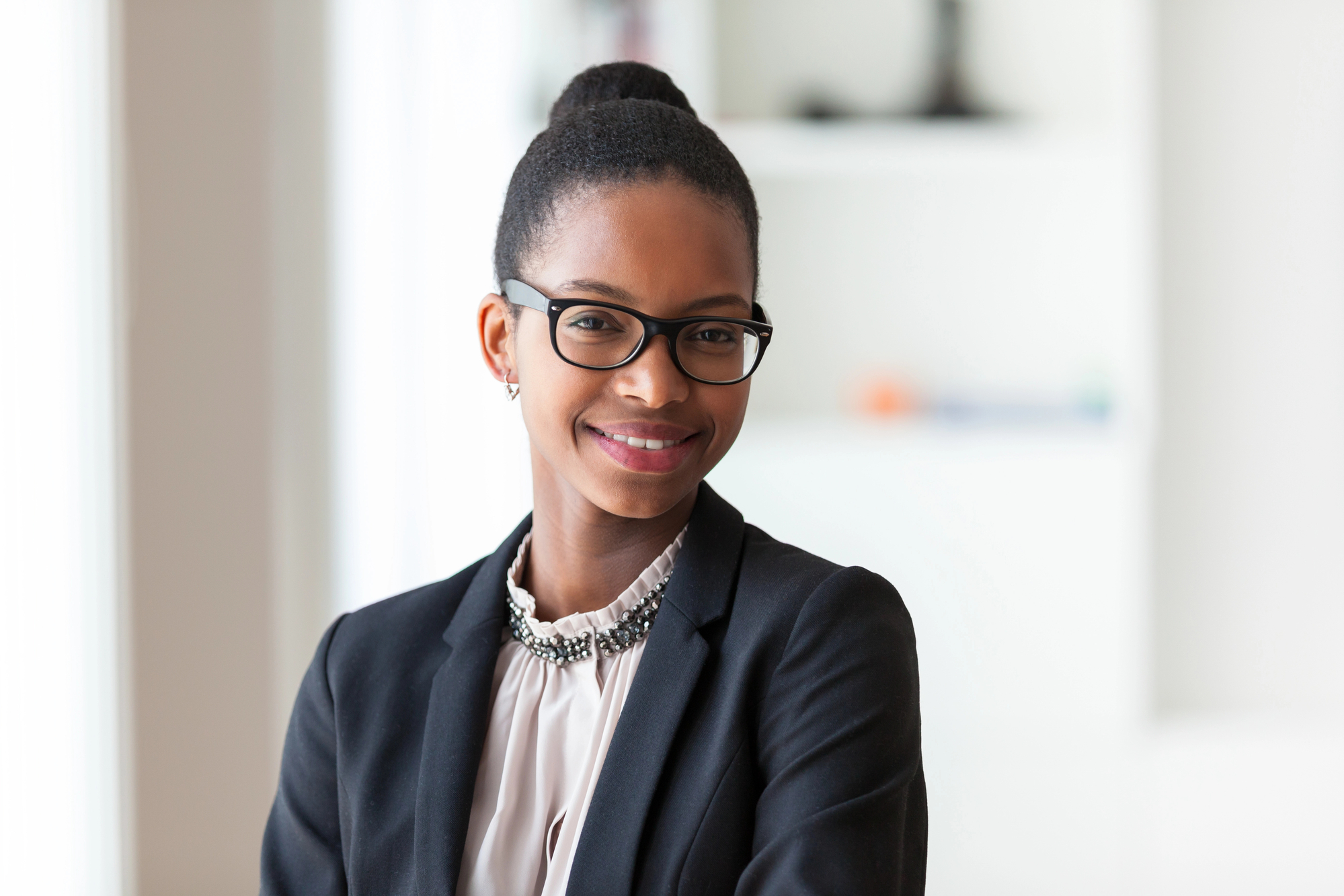 A person with dark hair pulled back into a bun, wearing glasses, a dark blazer, and a white blouse with a statement necklace, smiles confidently at the camera. The background is softly blurred, suggesting an office or professional setting.