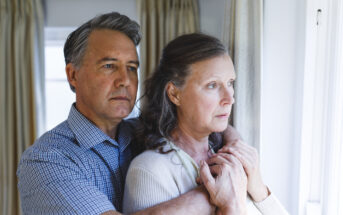 An older man and woman stand closely together near a window, looking outside with serious expressions. The man is behind the woman, holding her hands gently. Both have gray hair, and the room has light-colored curtains. The mood feels contemplative.