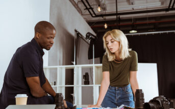 Two people, a man and a woman, are intently reviewing photographs on a table in a modern studio. Camera equipment, including lenses and a camera, is scattered on the table. The background features shelves, lighting equipment, and a minimalistic design.