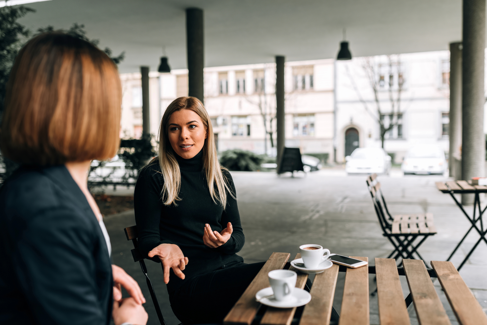 Two women are seated at an outdoor café, engaged in conversation. Both have cups of coffee and one woman's smartphone rests on the table. The background shows an open space with tall pillars, parked cars, and several building facades.