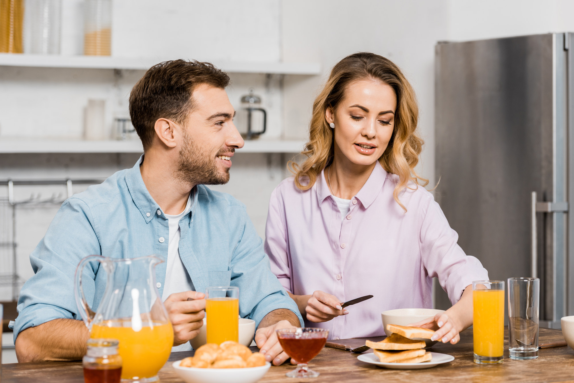 A man and woman are at a kitchen table enjoying breakfast. The woman is spreading jam on a piece of toast while the man, smiling, looks at her. Glasses of orange juice, a pitcher, croissants, and jars of jam and honey are on the table.