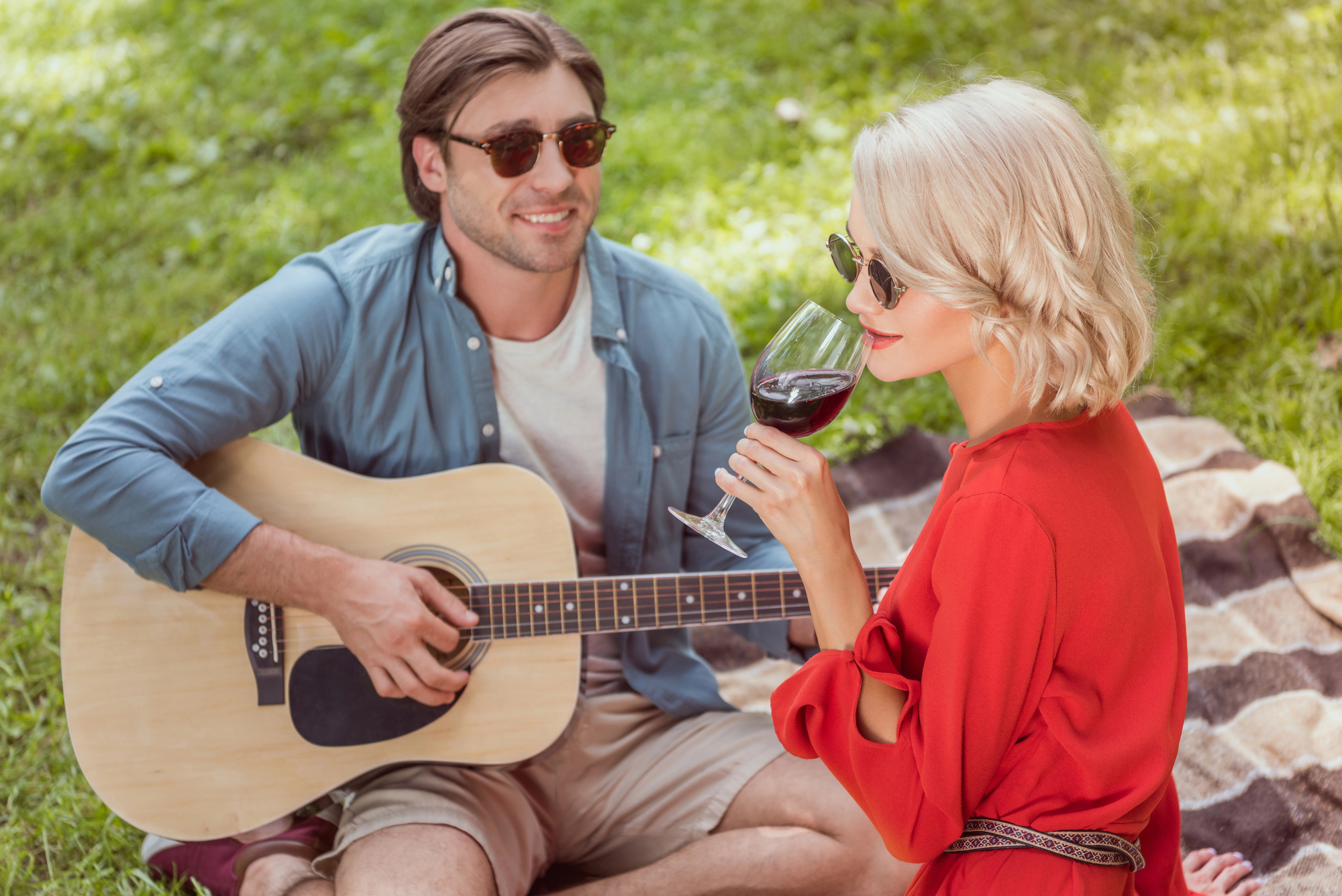 A man wearing sunglasses plays an acoustic guitar while sitting on a blanket in a park. A woman with short blonde hair and sunglasses, dressed in a red top, sits next to him, enjoying a glass of red wine. The scene is relaxed and sunlit.