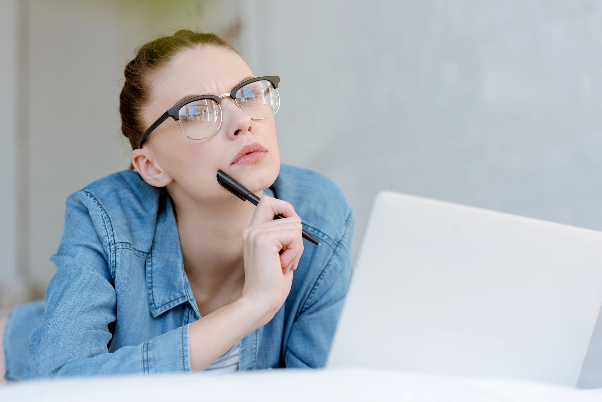 A person with glasses, wearing a denim jacket, is thoughtfully looking upwards while holding a pen under their chin. They are seated in front of an open laptop, appearing to be in deep thought or contemplation.