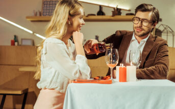 A man in glasses and a blazer pours wine into a woman's glass at a restaurant. The woman, with long blonde hair and wearing a white blouse, smiles while looking at him. They are seated at a table set with candles, wine glasses, and a white tablecloth.