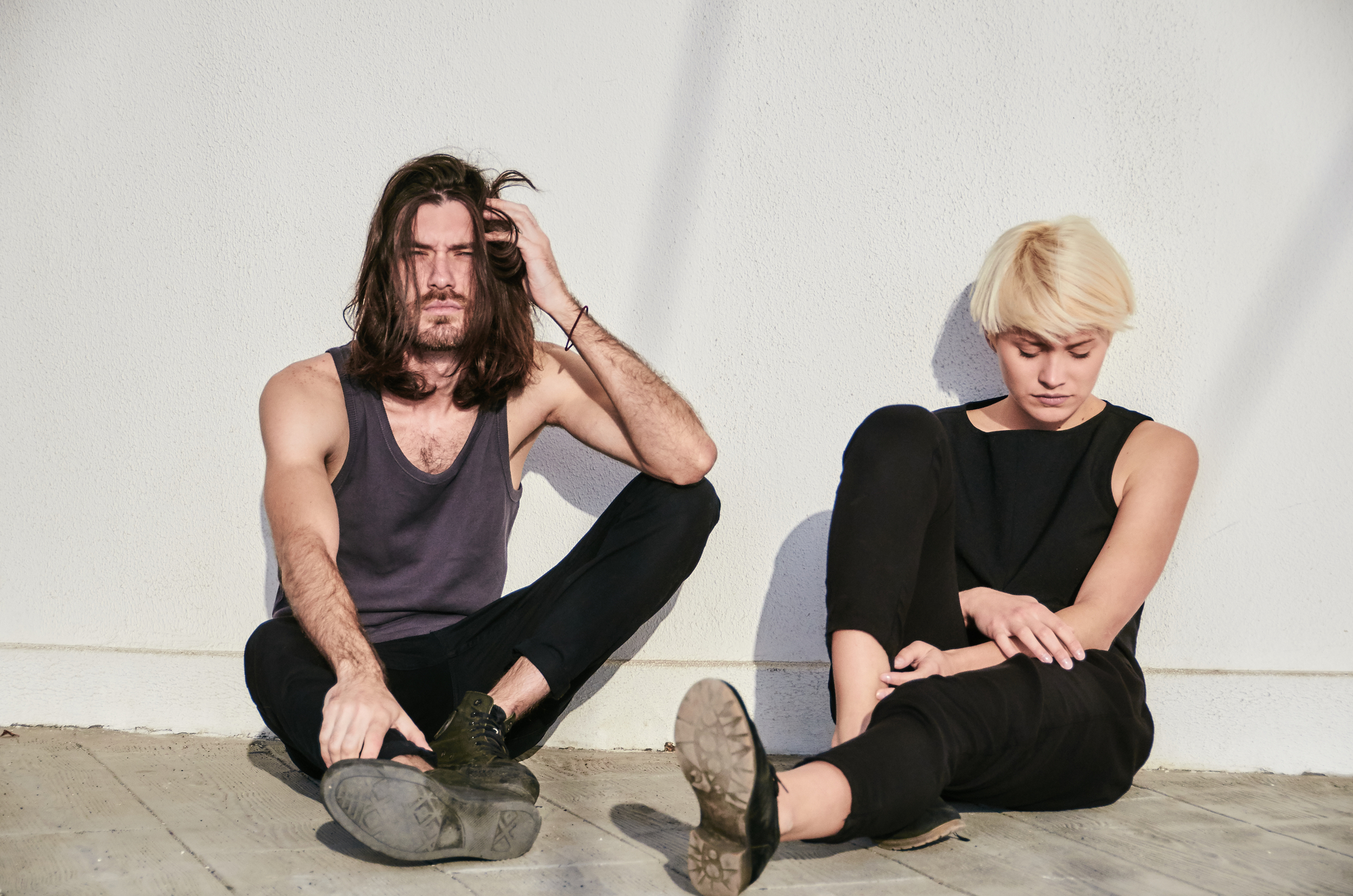 A man and a woman are sitting on the ground against a white wall. The man has long dark hair, a beard, and wears a black tank top and pants, resting one hand on his head. The woman has short blonde hair, wears a black sleeveless top and pants, looking down.