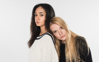 Two young women pose against a white background. The woman on the left has long dark hair and is wearing a white sweater, looking slightly to the right. The woman on the right has long blonde hair, is wearing a black sweater, and rests her head on the other woman's shoulder.