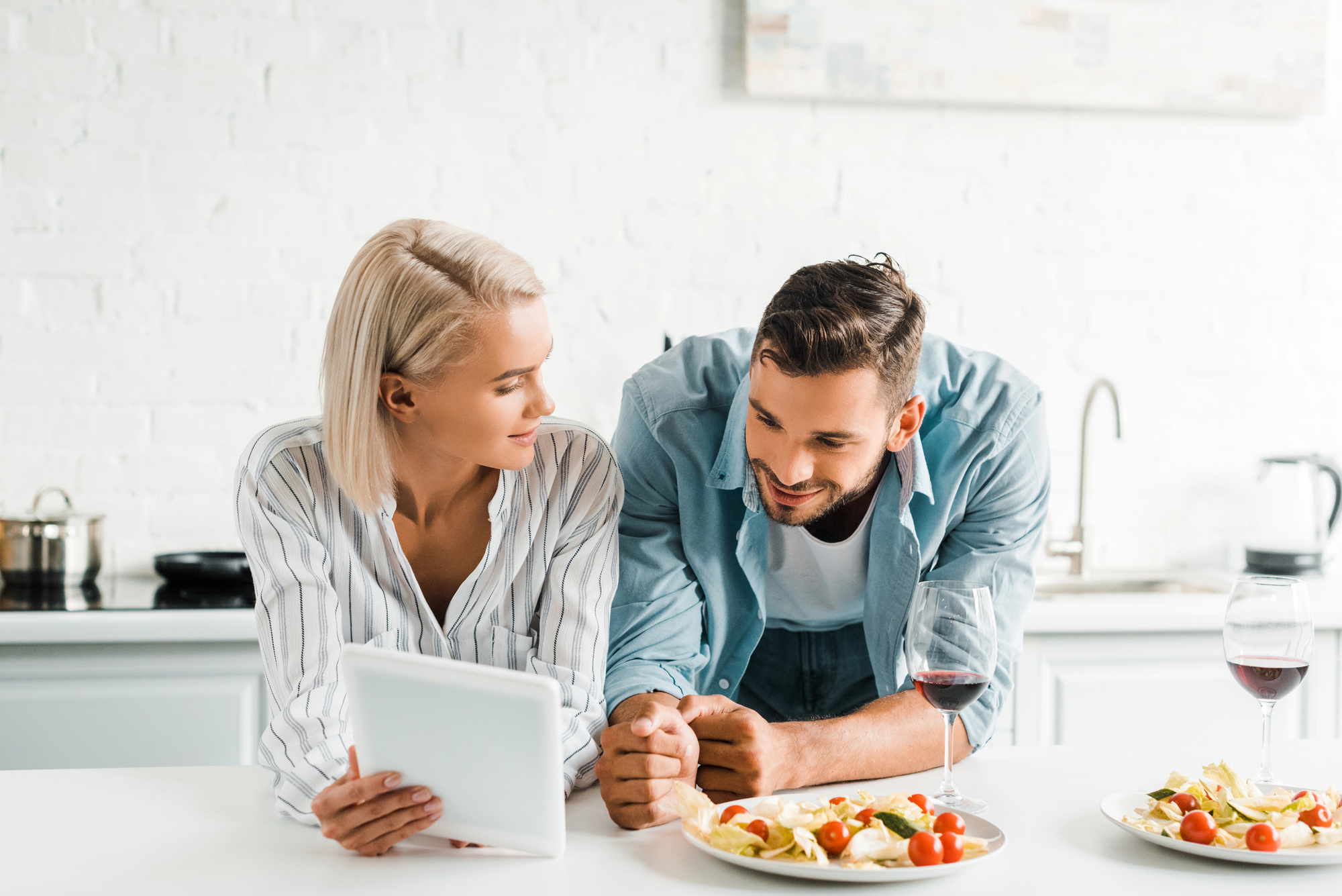 A couple is leaning on a kitchen counter, smiling and looking at each other. The woman holds a tablet, and two plates of food along with two glasses of red wine are placed on the counter. The kitchen background is bright and modern.