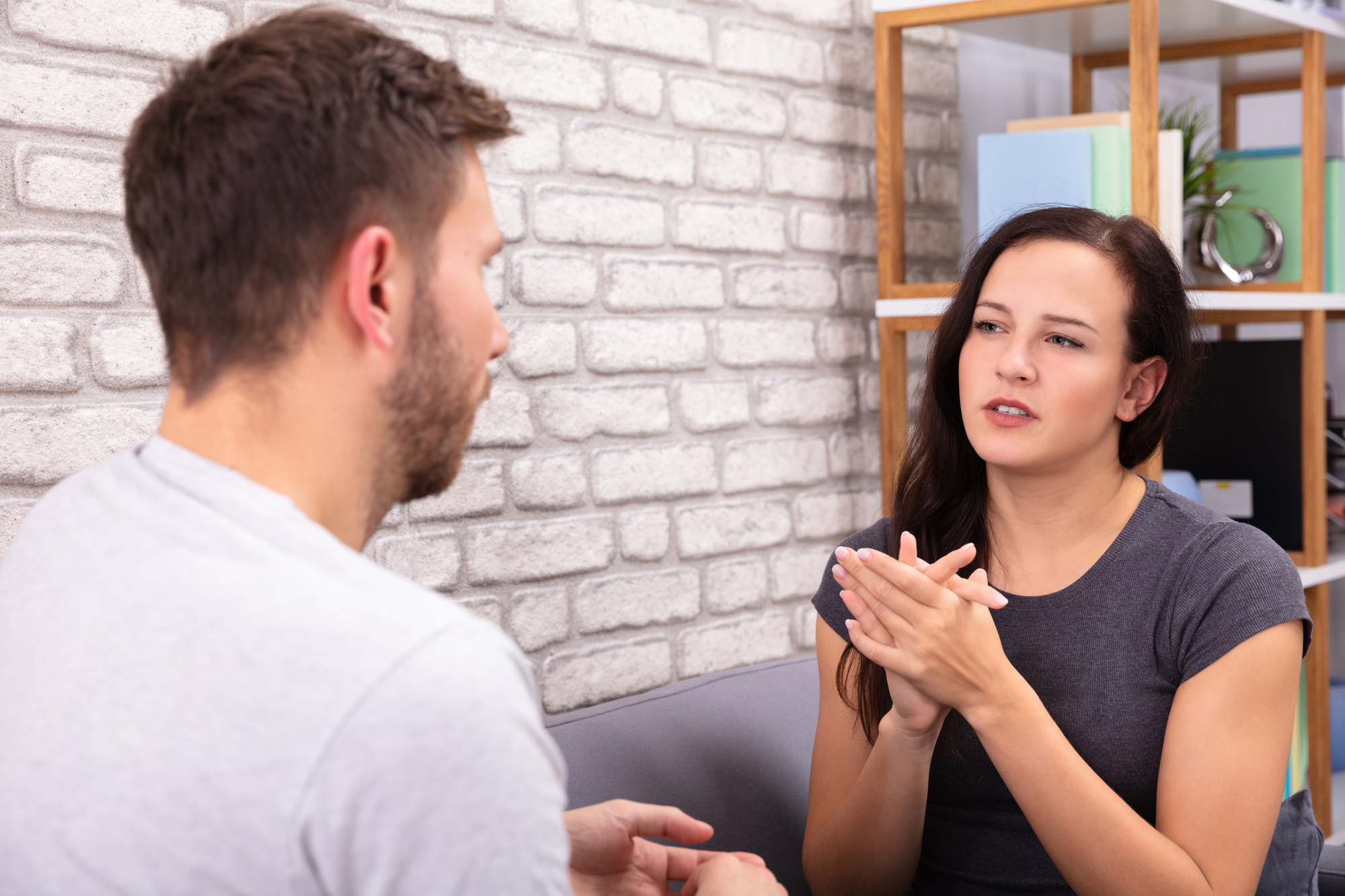 A woman with long dark hair is sitting on a couch, engaged in a conversation with a man who has short brown hair and a beard. She is gesturing with her hands, while the man listens intently. They are in a room with a white brick wall and a wooden shelving unit.