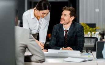 A man in a suit smiles and looks at a woman in a white blouse who is leaning over the table, pointing at a document. They are having a discussion in a modern office with a blurred background, including another person out of focus in the foreground.