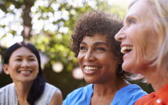 Three women are sitting outdoors, smiling and enjoying each other's company. They are in a relaxed, natural setting with tree foliage in the background, creating a bright and cheerful atmosphere. Each woman displays a warm, joyful expression.