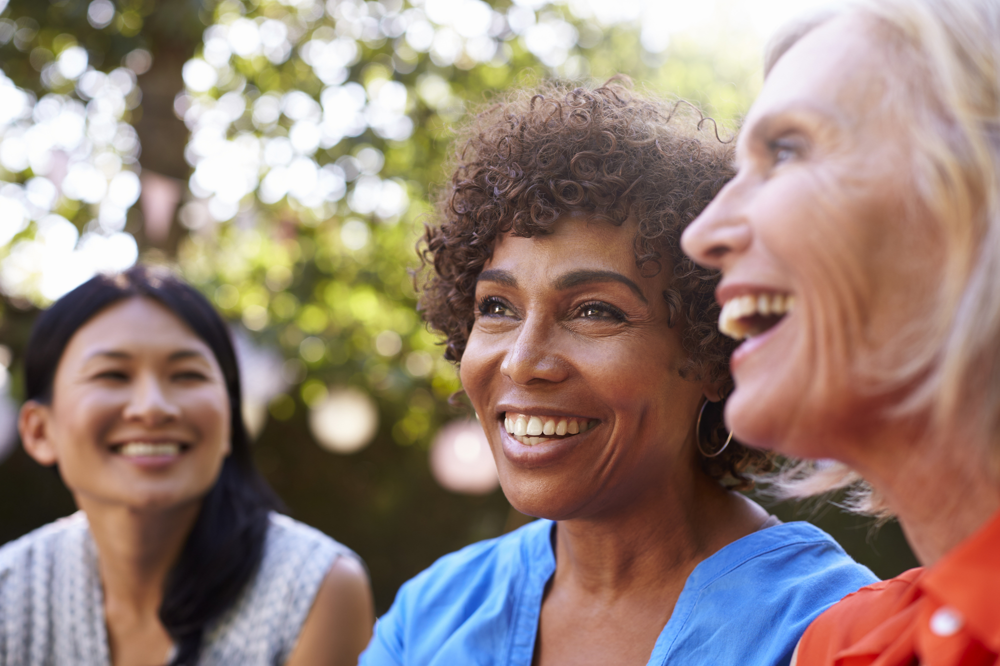 Three women are sitting outdoors, smiling and enjoying each other's company. They are in a relaxed, natural setting with tree foliage in the background, creating a bright and cheerful atmosphere. Each woman displays a warm, joyful expression.