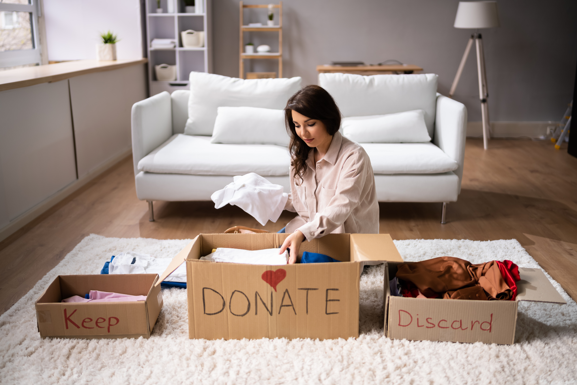 A woman sits on a beige carpet in a living room, sorting clothes into three boxes labeled "Keep," "Donate," and "Discard." The room has a white sofa, shelving, and a lamp in the background. She is holding a piece of clothing above the "Donate" box.