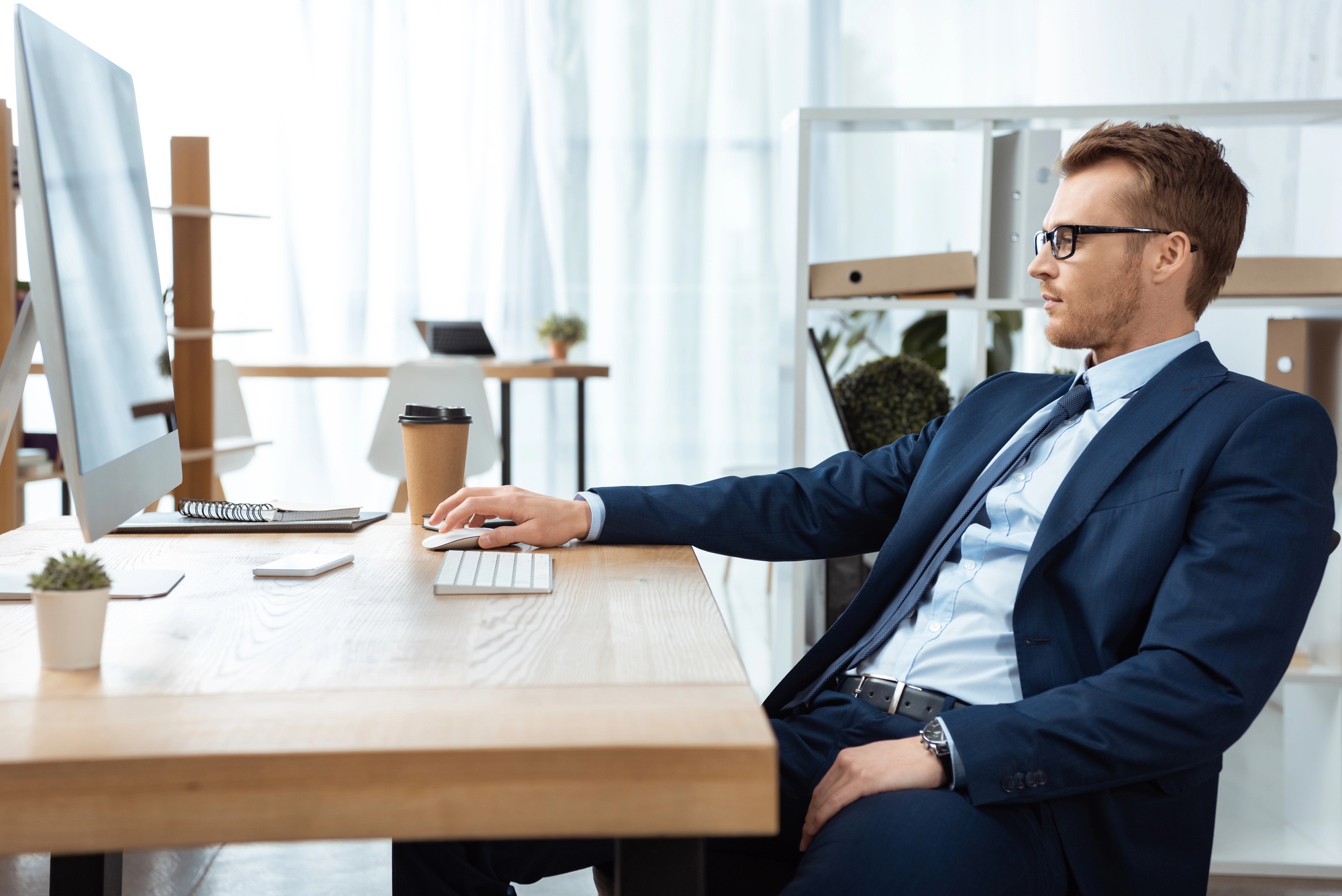 A man in a blue suit and glasses sits at a wooden desk, working on a desktop computer in a modern, bright office environment. A coffee cup, smartphone, and small potted plants are on the desk. Shelves and light curtains are visible in the background.