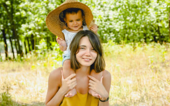 A woman with shoulder-length hair is giving a piggyback ride to a small child who is wearing a large straw hat. They are outdoors in a sunny, wooded area, and both are smiling happily at the camera. The background is lush with green trees and tall grass.