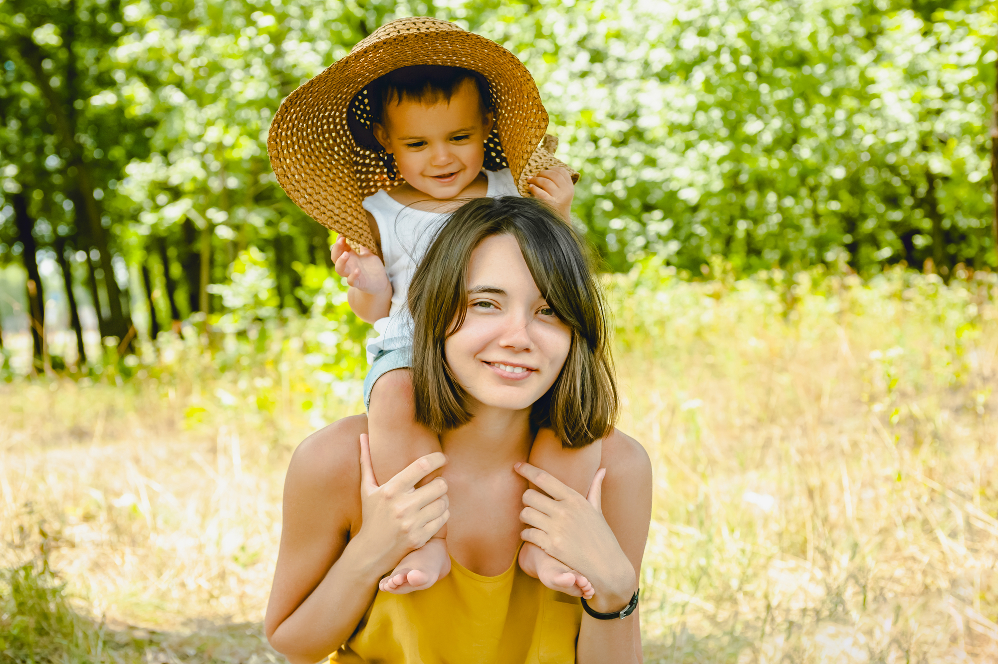 A woman with shoulder-length hair is giving a piggyback ride to a small child who is wearing a large straw hat. They are outdoors in a sunny, wooded area, and both are smiling happily at the camera. The background is lush with green trees and tall grass.