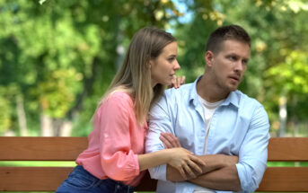 A woman with a concerned expression leans towards a man with folded arms, both seated on a park bench. She appears to be talking or comforting him, while he looks away thoughtfully. Green trees and foliage are visible in the blurred background.