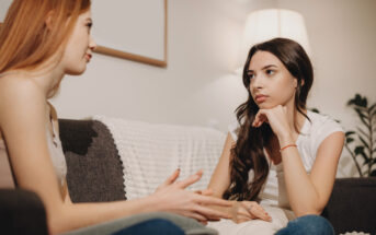 Two young women are sitting on a couch in a well-lit living room. One with long brown hair listens attentively with her chin resting on her hand, while the other with long red hair speaks, gesturing with her hands. They appear to be engaged in a serious conversation.