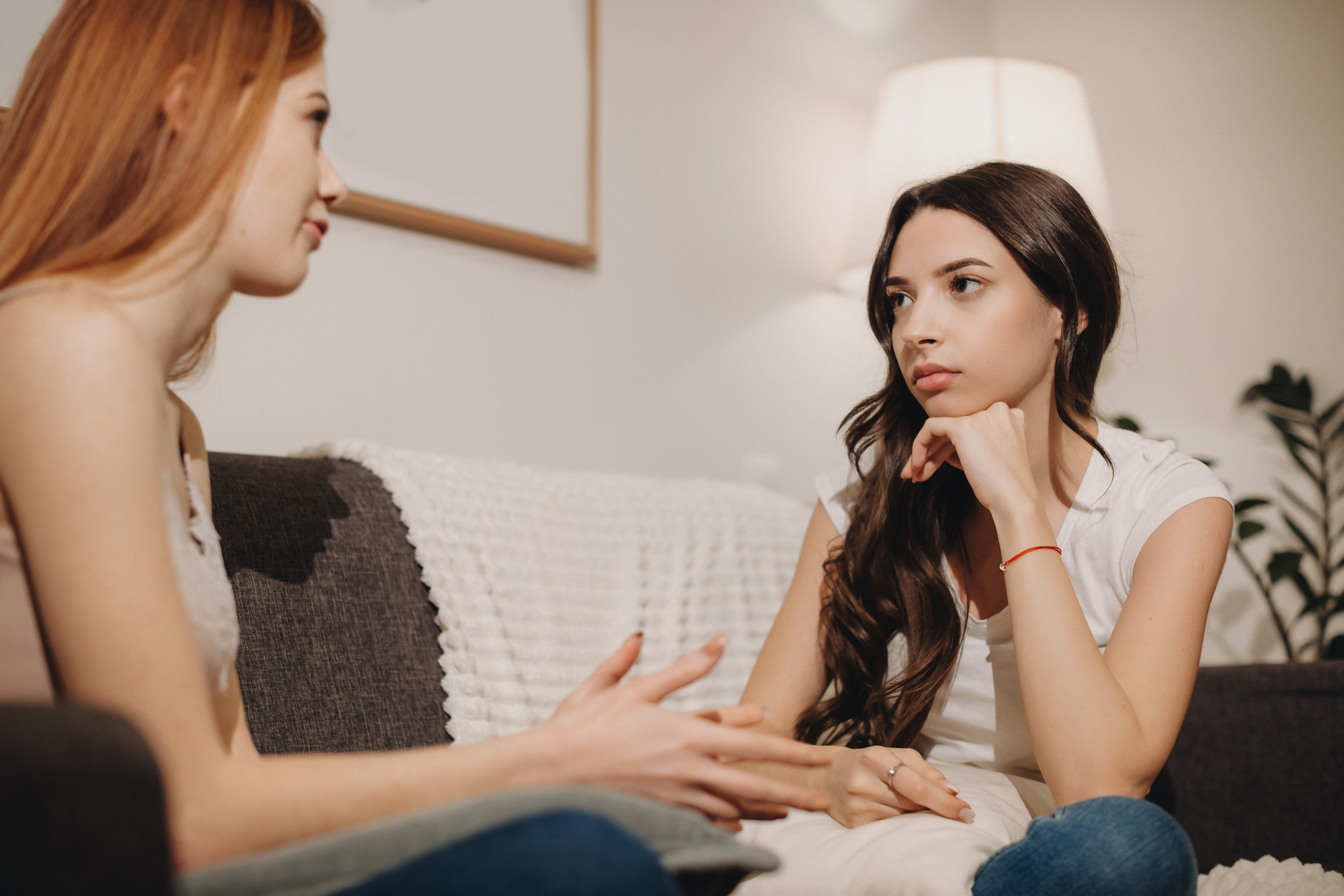 Two young women are sitting on a couch in a well-lit living room. One with long brown hair listens attentively with her chin resting on her hand, while the other with long red hair speaks, gesturing with her hands. They appear to be engaged in a serious conversation.