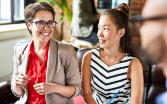 Two women are seated and smiling in an office setting. The woman on the left wearing glasses and a beige blazer over a red top, holds a pen. The woman on the right has long hair tied back and is dressed in a black-and-white striped dress, also holding a pen.