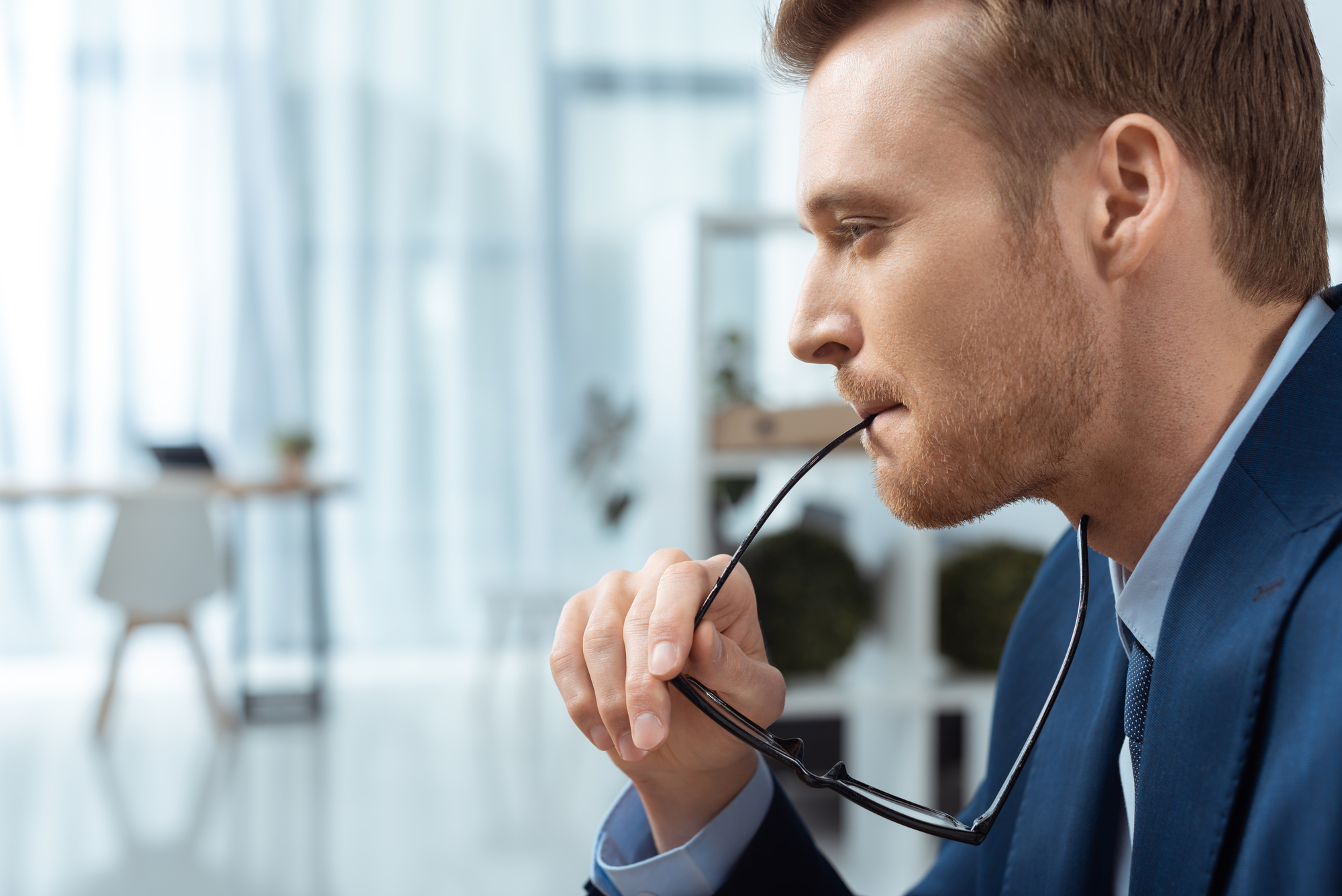 Side profile of a man in a blue suit deep in thought, holding eyeglasses to his mouth. The blurred background features an airy, modern office space with light-colored furnishings and large windows, allowing natural light to fill the room.