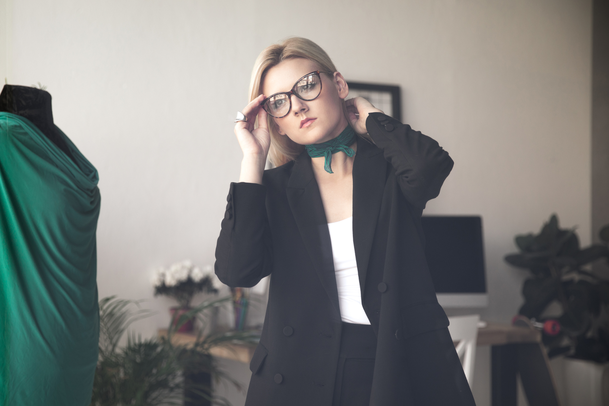 A person with blonde hair and glasses wearing a black blazer over a white top adjusts their glasses. They are standing in a room with various objects like a computer, plants, and a dress form with a green garment.