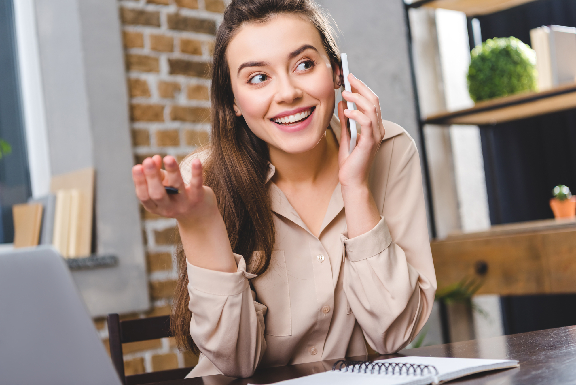 A woman with long brown hair is smiling while talking on a phone in an office setting. She gestures with her left hand and has a notebook and laptop in front of her on the desk. The background features a brick wall and a shelf with decorative items.