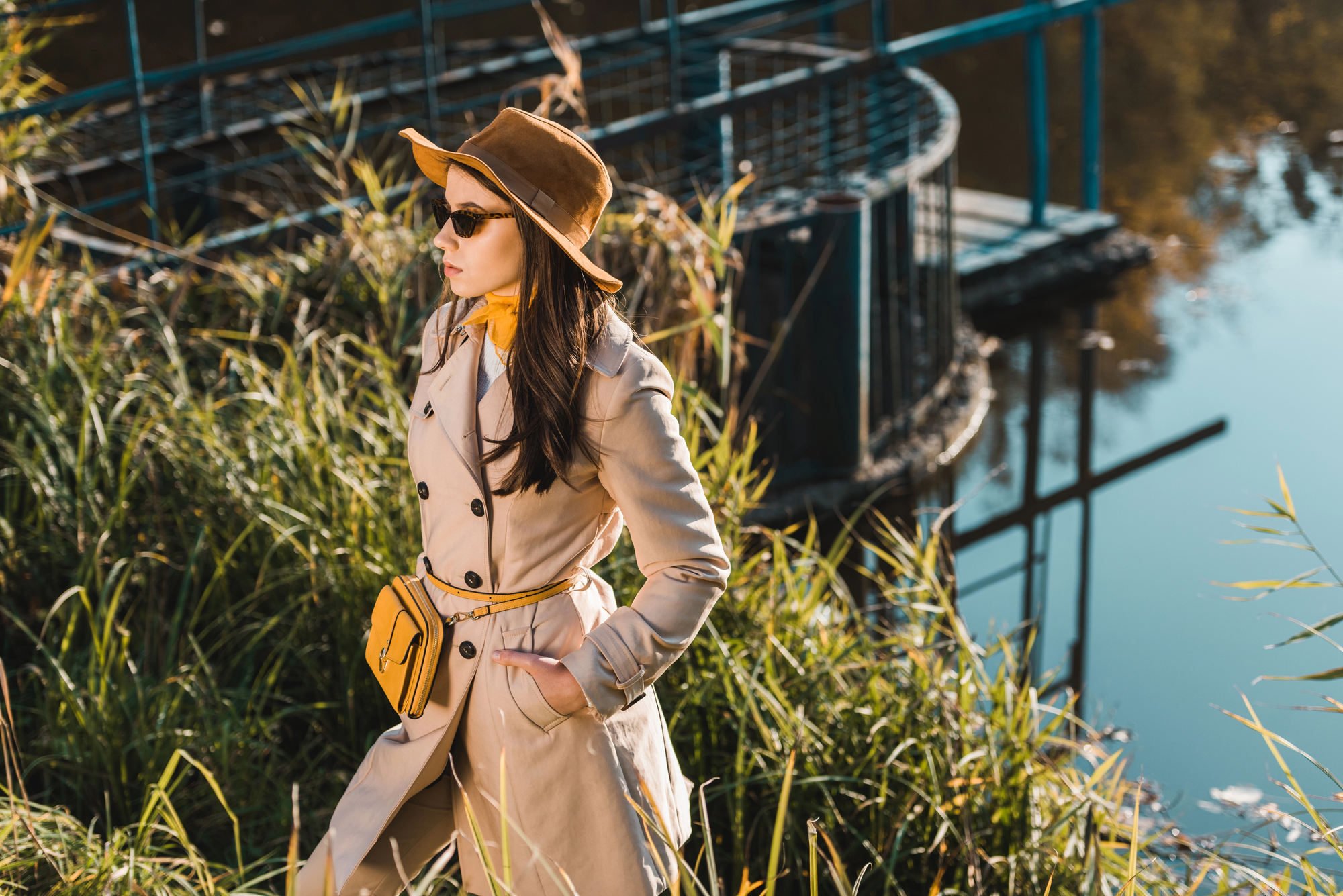 A woman in a beige trench coat and wide-brimmed hat stands near a body of water surrounded by tall grass. She wears sunglasses and a yellow crossbody bag, looking to the side. A structure with blue railings is partially visible behind her against a clear, reflecting sky.