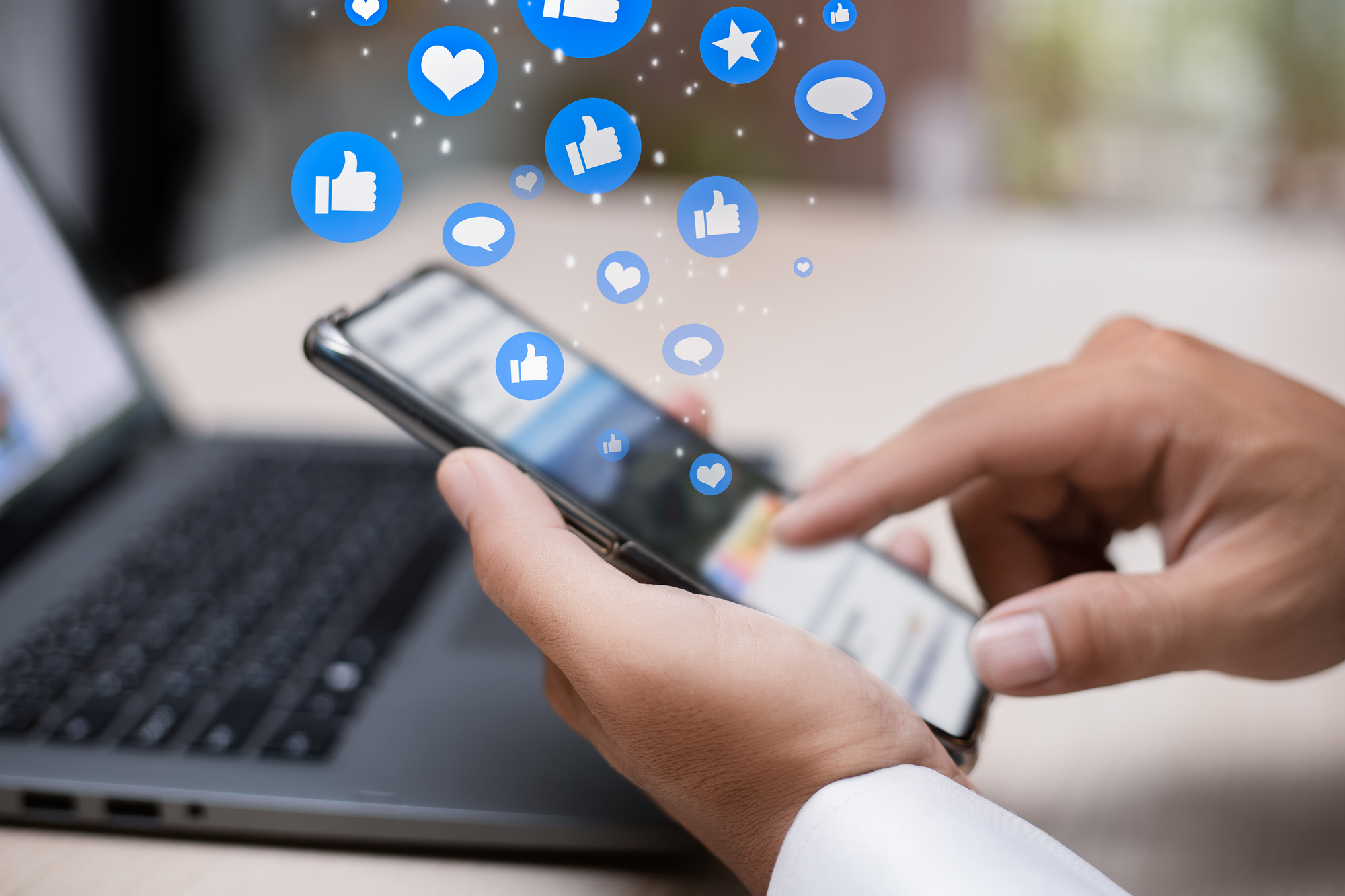 A person using a smartphone with a laptop in the background. Blue icons representing social media interactions such as likes, comments, and shares float above the phone screen, illustrating online engagement and connectivity.