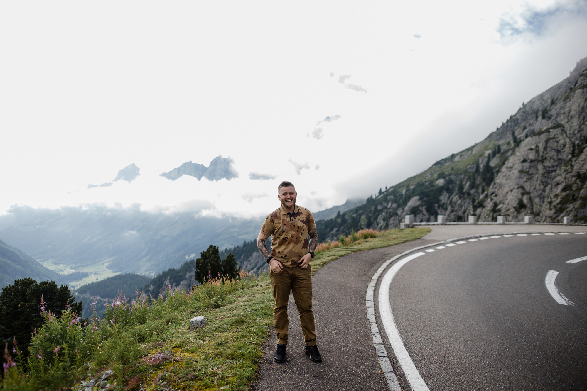 A person in a brown camouflage shirt and khaki pants stands smiling on a mountain road with a white dashed line. In the background, the scene features towering mountains partially covered by clouds and lush green valleys. Flowers and trees line the hillside.