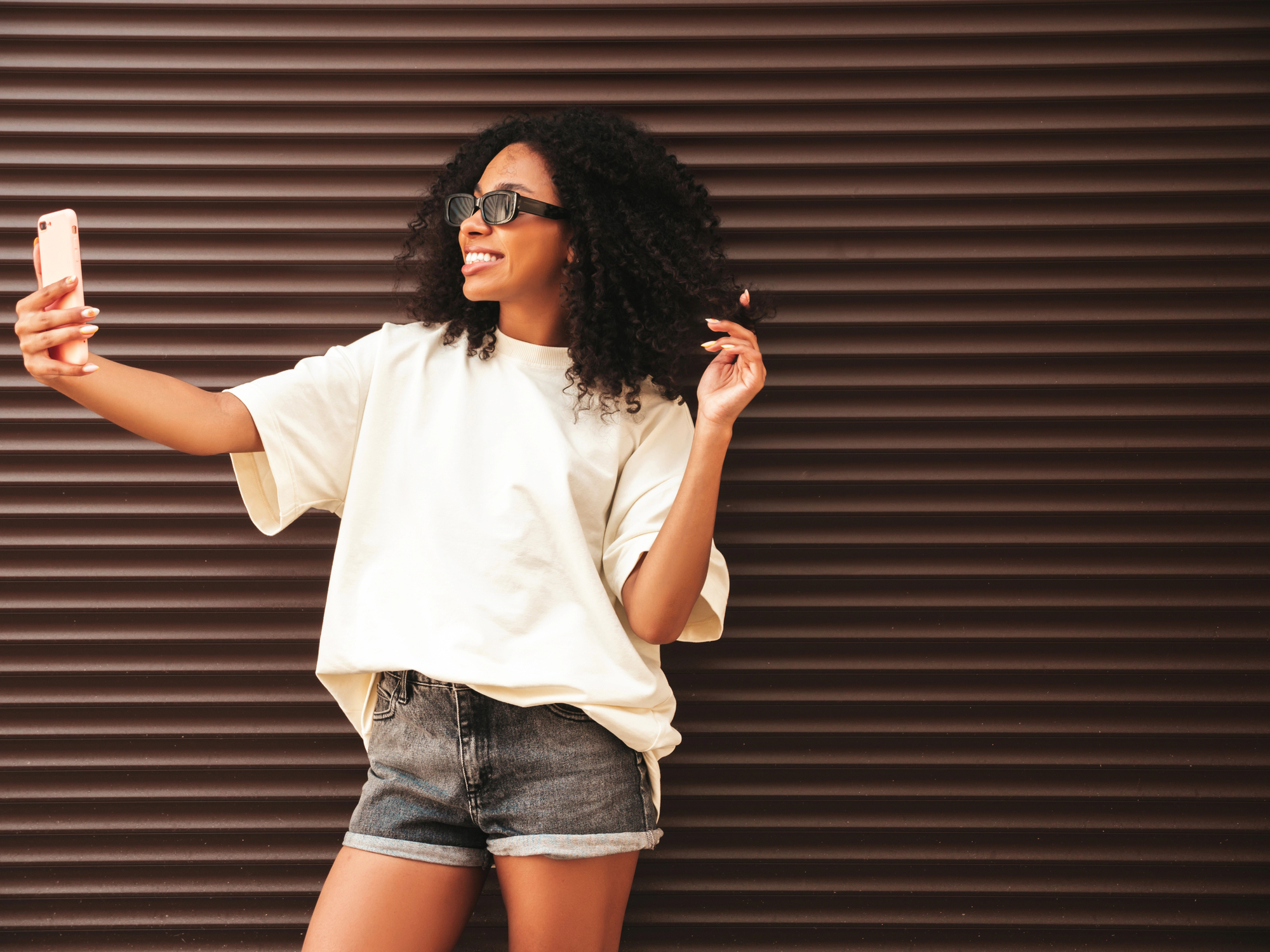 A woman with curly hair wearing sunglasses, an oversized white T-shirt, and denim shorts poses for a selfie with a smartphone. She is smiling and standing against a dark brown corrugated metal background.