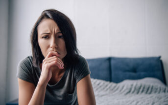 A woman with short dark hair sits on the edge of a bed in a pensive, worried expression. She is resting her chin on her hand and looking downward. She is wearing a gray t-shirt, and the bed behind her is covered with a textured gray blanket.