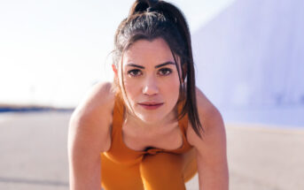 A woman is in a forward-leaning position, as if about to start a race, with her hair tied back in a high ponytail. She is wearing an orange athletic outfit and looking intently at the camera. The background features a blurred blue structure and pavement.