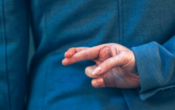 A person wearing a blue jacket is holding their right hand behind their back with their fingers crossed. The gesture is traditionally associated with superstition or deceit. The focus is on the hand and the texture of the jacket.