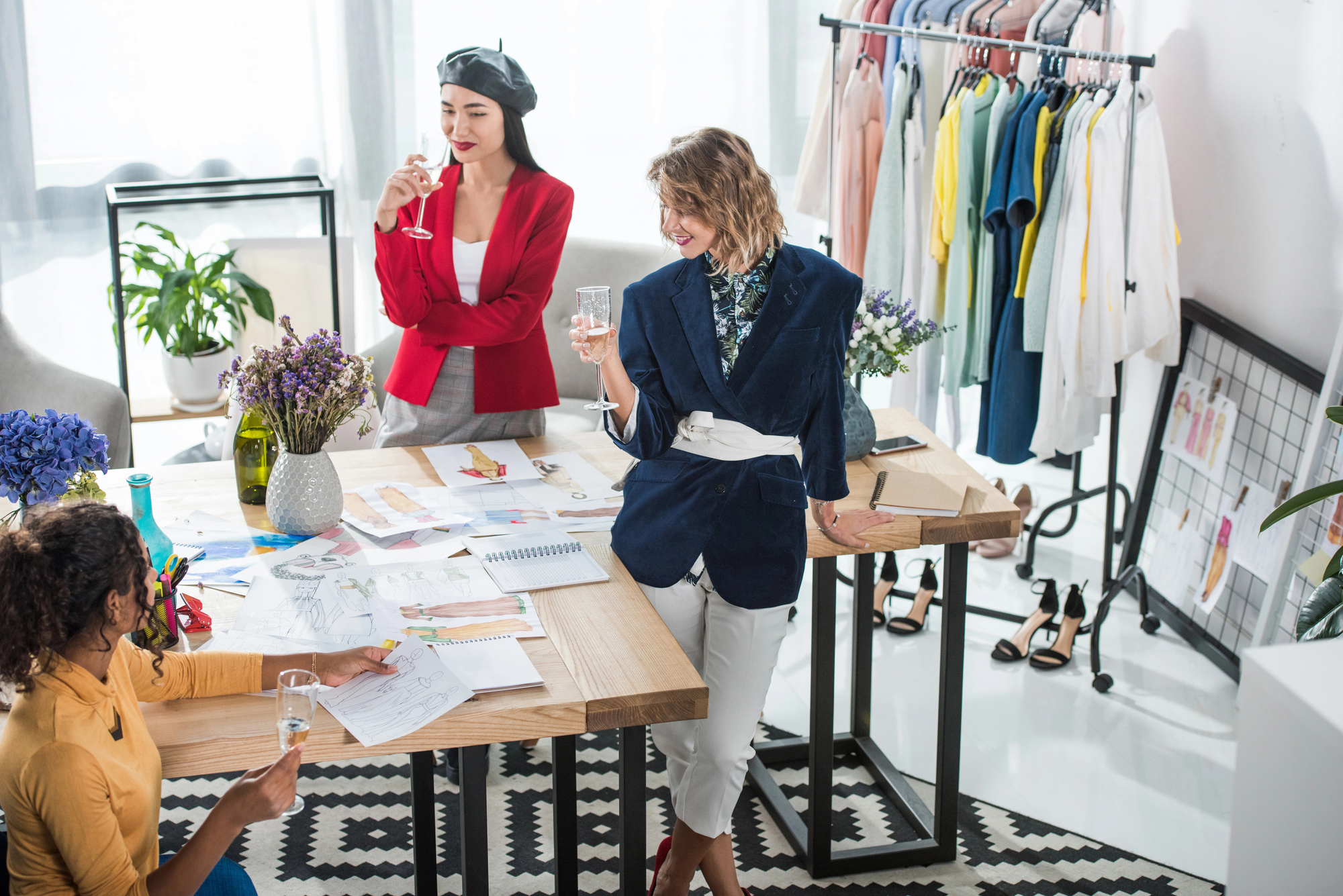 Three women in a modern studio engage in a lively discussion. One leans against the table holding a glass, another sits while pointing at papers, and the third stands with a drink. The workspace is adorned with fashion sketches, fabric swatches, colorful clothes racks, and flowers.