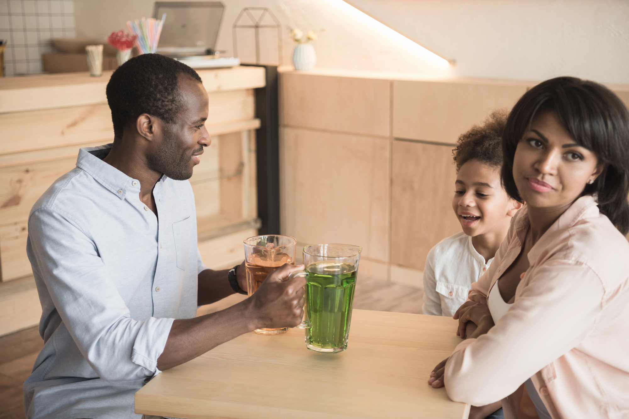 A man, woman, and child are seated at a wooden table in a modern, cozy café. The man and child are engaged in conversation, while the woman looks off-camera and holds a drink. Two large pitchers, one with green liquid and one with orange liquid, are on the table.