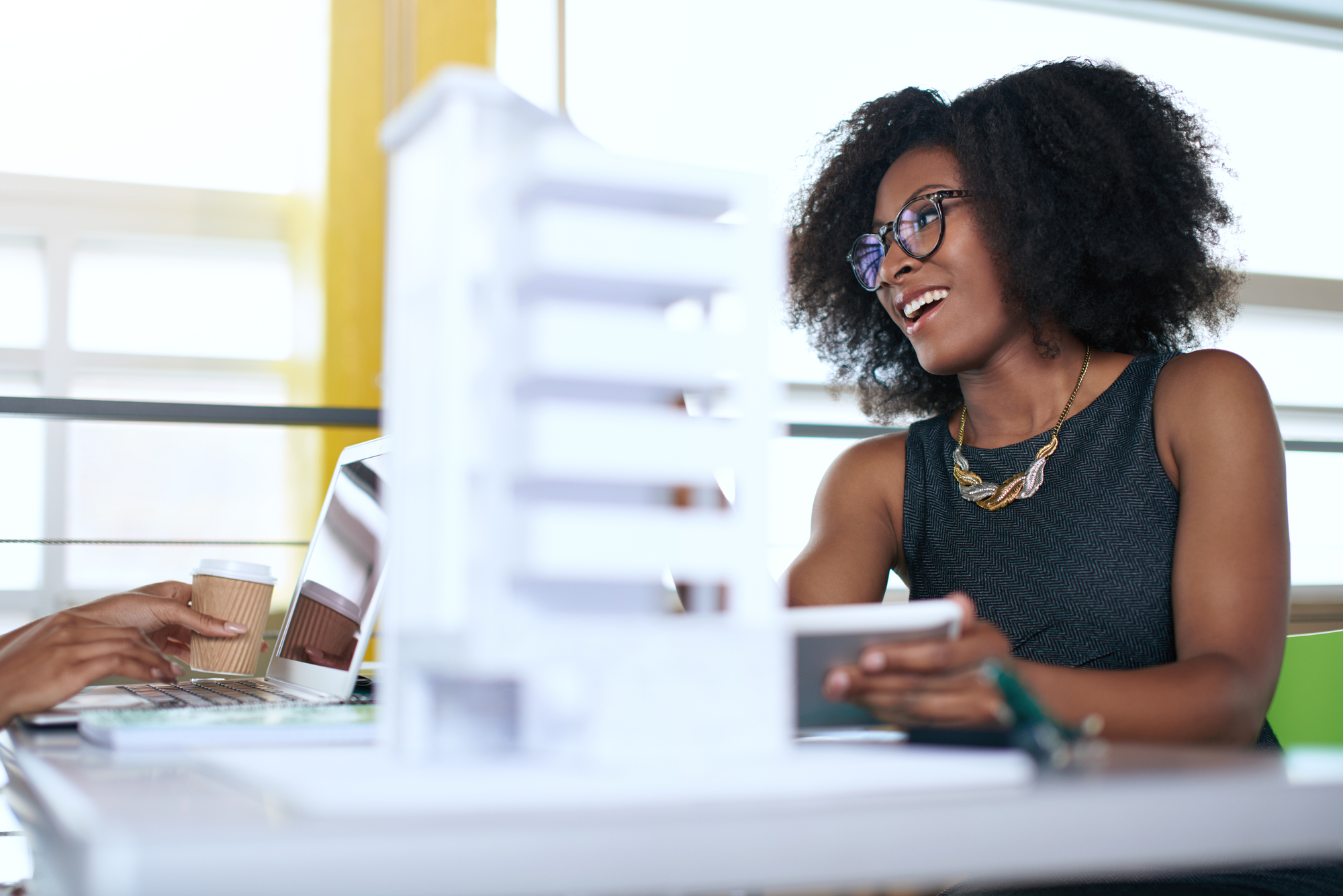 A woman with curly hair and glasses, wearing a dark sleeveless top and a necklace, smiles while sitting at a desk. She appears engaged in conversation. In front of her is a white architectural model. Another person’s hand holding a coffee cup is partially visible.