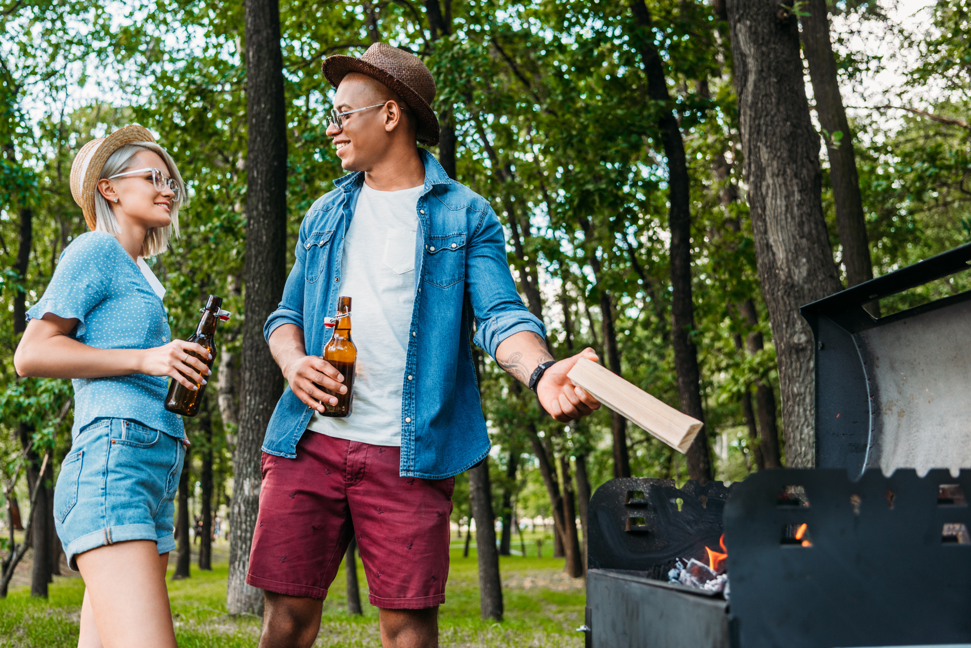 Two people stand outdoors near a grill in a park. One person, wearing a hat and sunglasses, holds a beer bottle and wood piece, while the other, also in a hat and sunglasses, holds a beer bottle. Trees and greenery are visible in the background.