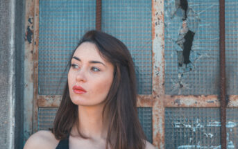 A woman with long brown hair and red lipstick is standing in front of a weathered, broken window with a metal grid. She is looking to her left, with a thoughtful expression. The background shows signs of decay and rust, creating a contrast with her appearance.