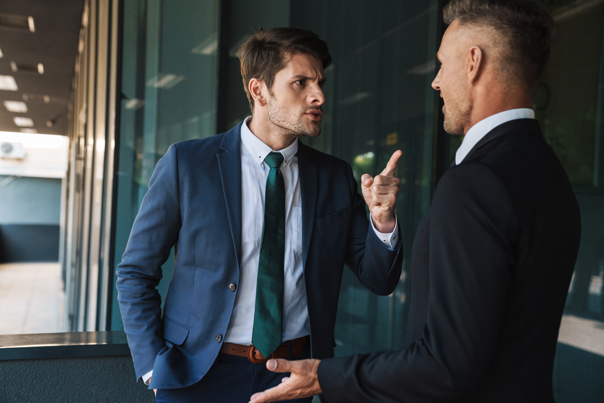 Two men in business attire engaging in a heated discussion. The man on the left, wearing a blue suit and green tie, points his finger at the other man, who is dressed in a black suit. They stand in a hallway with glass walls.