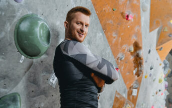 A person with red hair and a beard is standing with crossed arms in front of a rock climbing wall. They are wearing a black and grey geometric-patterned long-sleeve shirt and appear to be smiling confidently. Various climbing holds are visible on the wall.