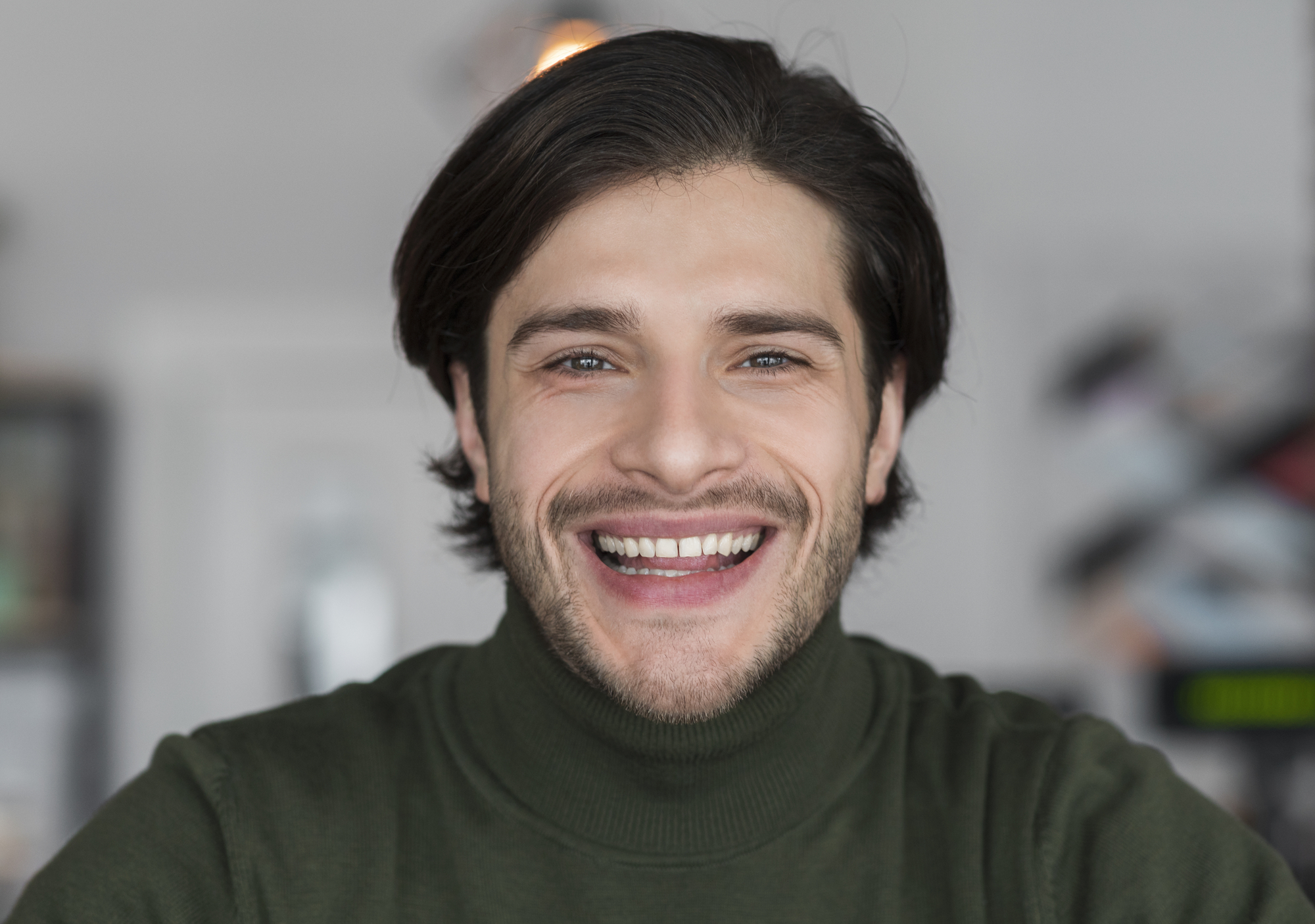 A man with medium-length dark hair and a beard is smiling warmly at the camera. He is wearing a dark green turtleneck sweater. The background is blurred, with soft lighting creating a cozy atmosphere.