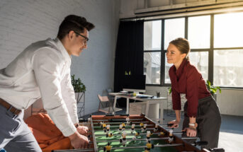 Two office workers, a man in a white shirt and a woman in a red blouse, play foosball in a brightly lit office space with large windows. The room features modern decor with a computer desk and plants in the background. They are smiling and enjoying the game.