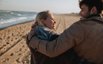 A couple wearing jackets embraces and smiles at each other while walking along a sandy beach. The ocean waves gently lap the shore, and tire tracks are visible in the sand. The sky is clear, indicating a calm and pleasant day.