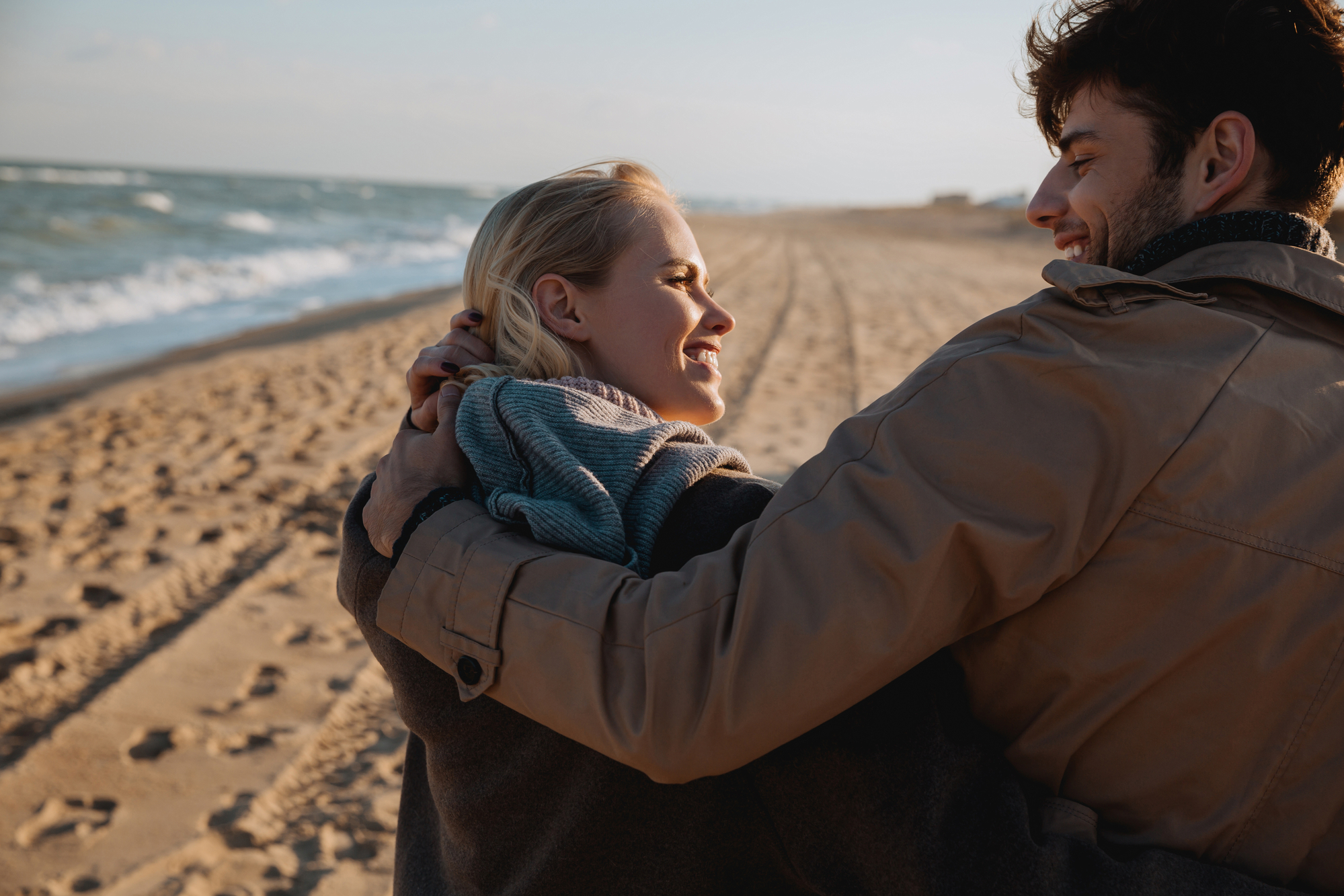 A couple wearing jackets embraces and smiles at each other while walking along a sandy beach. The ocean waves gently lap the shore, and tire tracks are visible in the sand. The sky is clear, indicating a calm and pleasant day.