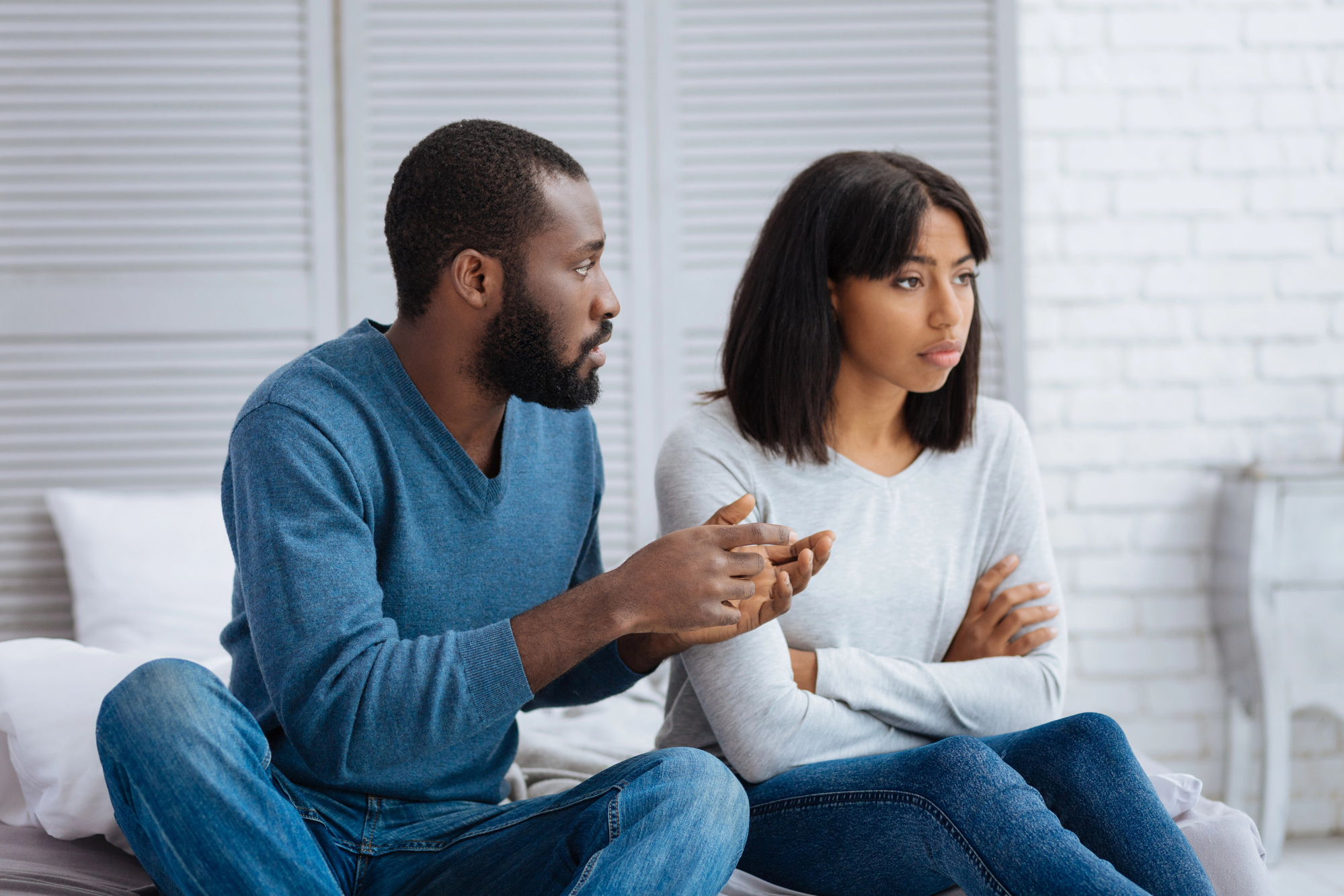 A man and woman sit on a bed in a serious conversation. The man, wearing a blue sweater and jeans, gestures with his hands while the woman, in a light grey sweater and jeans, has her arms folded, looking away with a concerned expression.