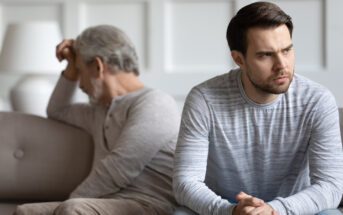 A younger man sits on a couch looking troubled, resting his hands together, while an older man in the background, also looking distressed, sits with his back to him, holding his head. Both men are wearing casual, light-colored clothing.