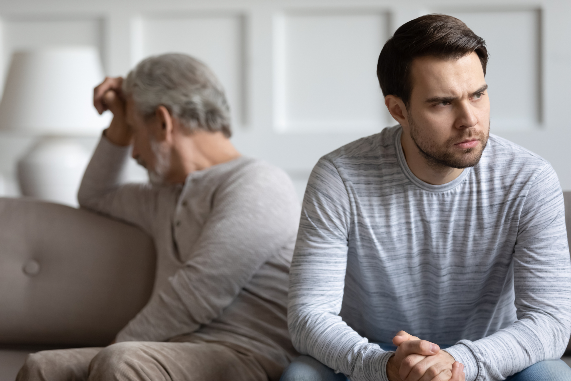 A younger man sits on a couch looking troubled, resting his hands together, while an older man in the background, also looking distressed, sits with his back to him, holding his head. Both men are wearing casual, light-colored clothing.