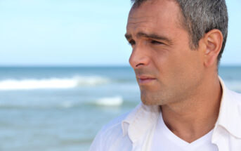 A man with short gray hair, wearing a white shirt over a white T-shirt, is standing on a beach. He is looking pensively into the distance with the sea and sky in the background.